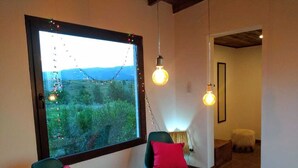 A cozy room with a large window, two chairs with red cushions, a wooden bench, and hanging light bulbs. - Kleines Landhaus in 25 Ha in den Bergen von Córdoba, im Herz Argentiniens (Villa Berna)