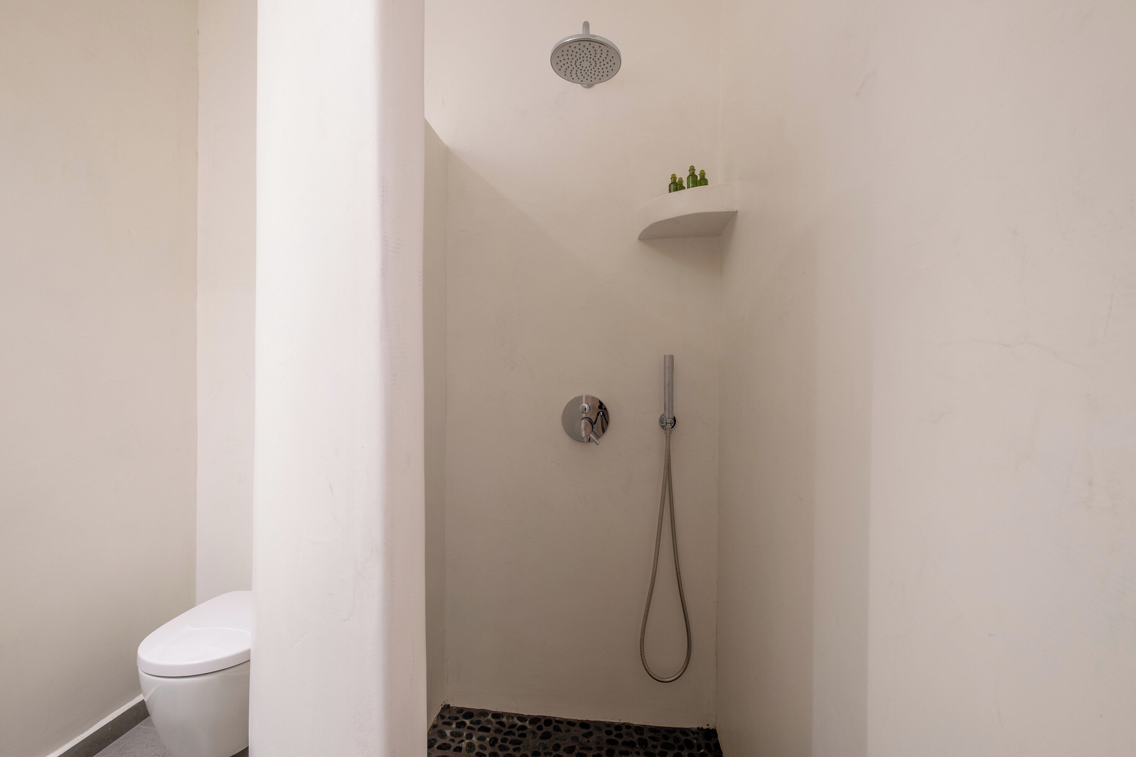 Une petite salle de bain blanche, équipée d’une douche, de toilettes et d’une étagère où sont disposés des objets décoratifs.