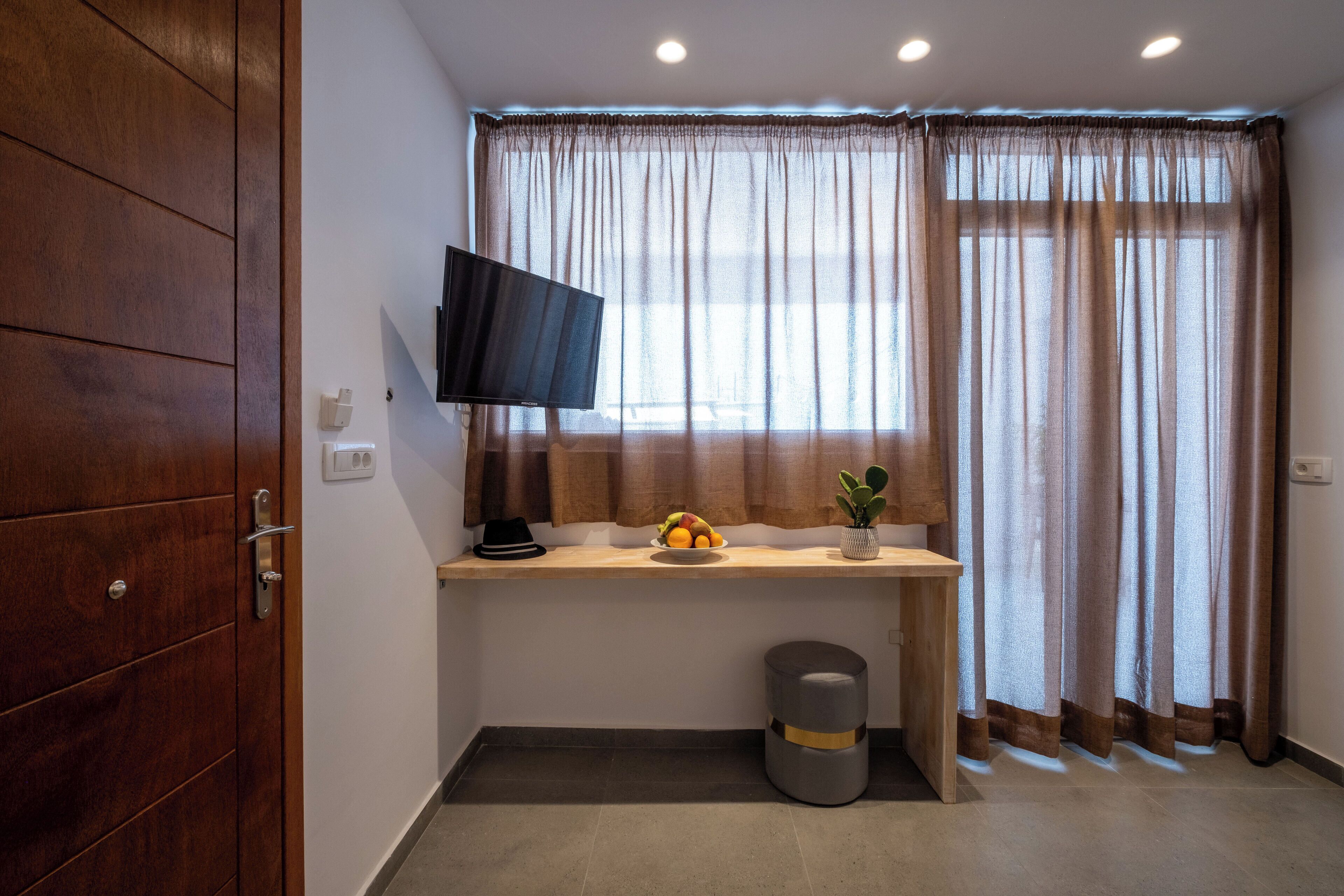 Une pièce comprenant une armoire en bois, un téléviseur à écran plat fixé au mur, un bureau avec un panier de fruits, une plante en pot et une poubelle grise.