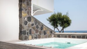 A stone wall with a white structure above it, a pool with clear water, and a view of the sea with trees in the background. - Sand Gem - Beach Apartments (Santorini)