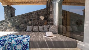 A sunlit room with a stone wall, a cushioned seating area, and a large window. - Sand Gem - Beach Apartments (Santorini)