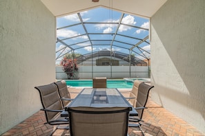 Un porche cubierto con una mesa y sillas, vista a una piscina y una casa al fondo.