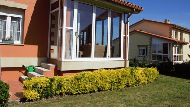 Ein Haus mit überdachter Veranda, einem Fenster mit Blick und einem kleinen Garten mit gelben Blumen.