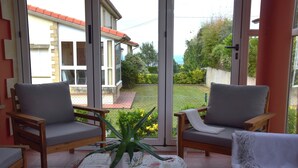 Eine sonnendurchflutete Terrasse mit zwei Holzstühlen, einem Tisch mit einer Pflanze und Blick auf ein Haus und Garten durch große Glasüren.