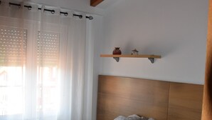 Ein Schlafzimmer mit einem Holz-Kopfteil, einem Bett mit grauer Bettwäsche, einem Fenster mit weißen Vorhängen und einem Regal mit zwei Dekorationen.