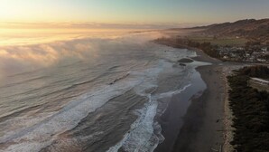 Eine Luftaufnahme einer Küstenlandschaft mit Strand, Wellen und einer Stadt im Hintergrund.
