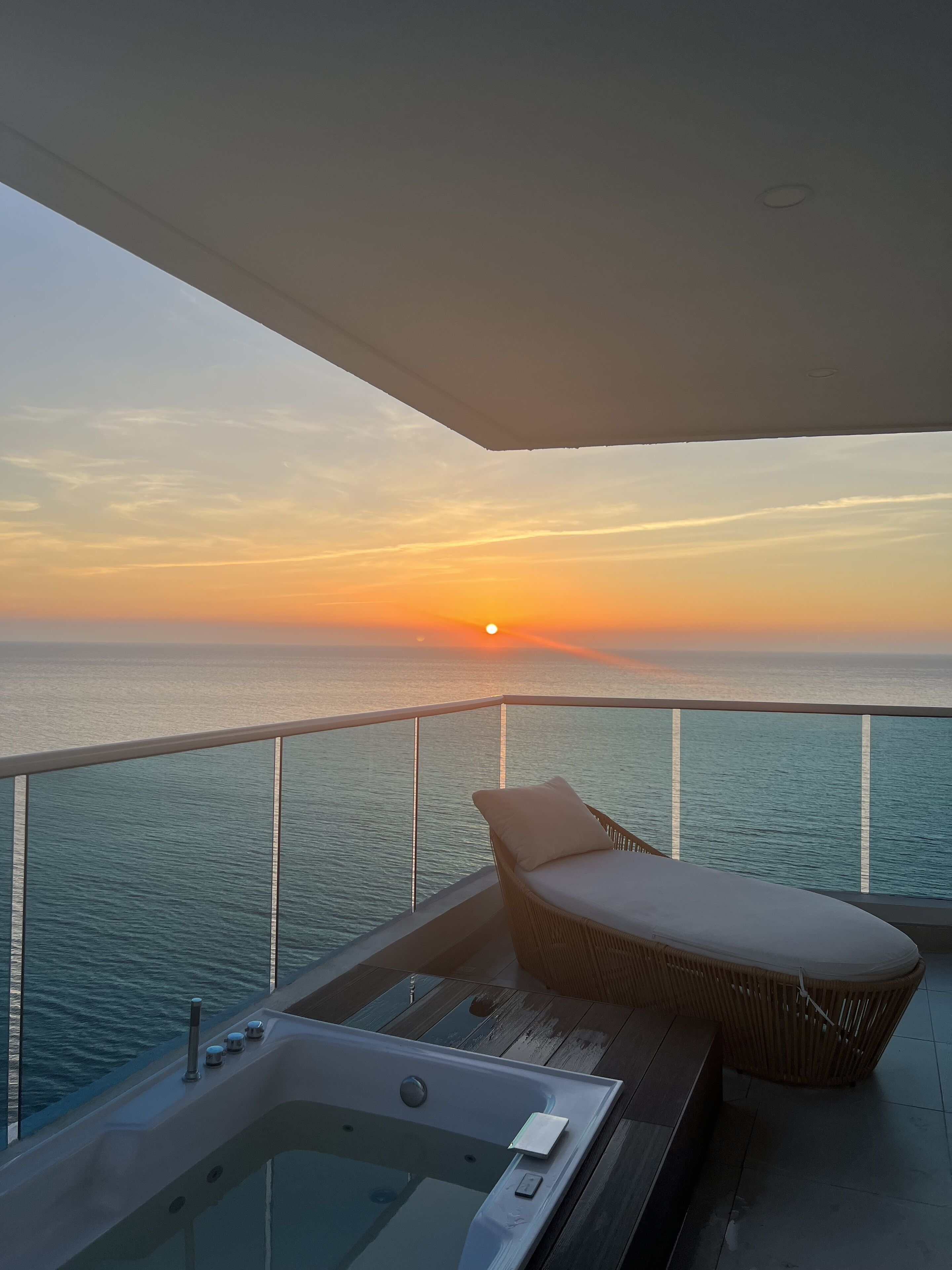 Uma varanda com vista para o pôr do sol sobre o oceano, uma cadeira de descanso e uma banheira de hidromassagem.