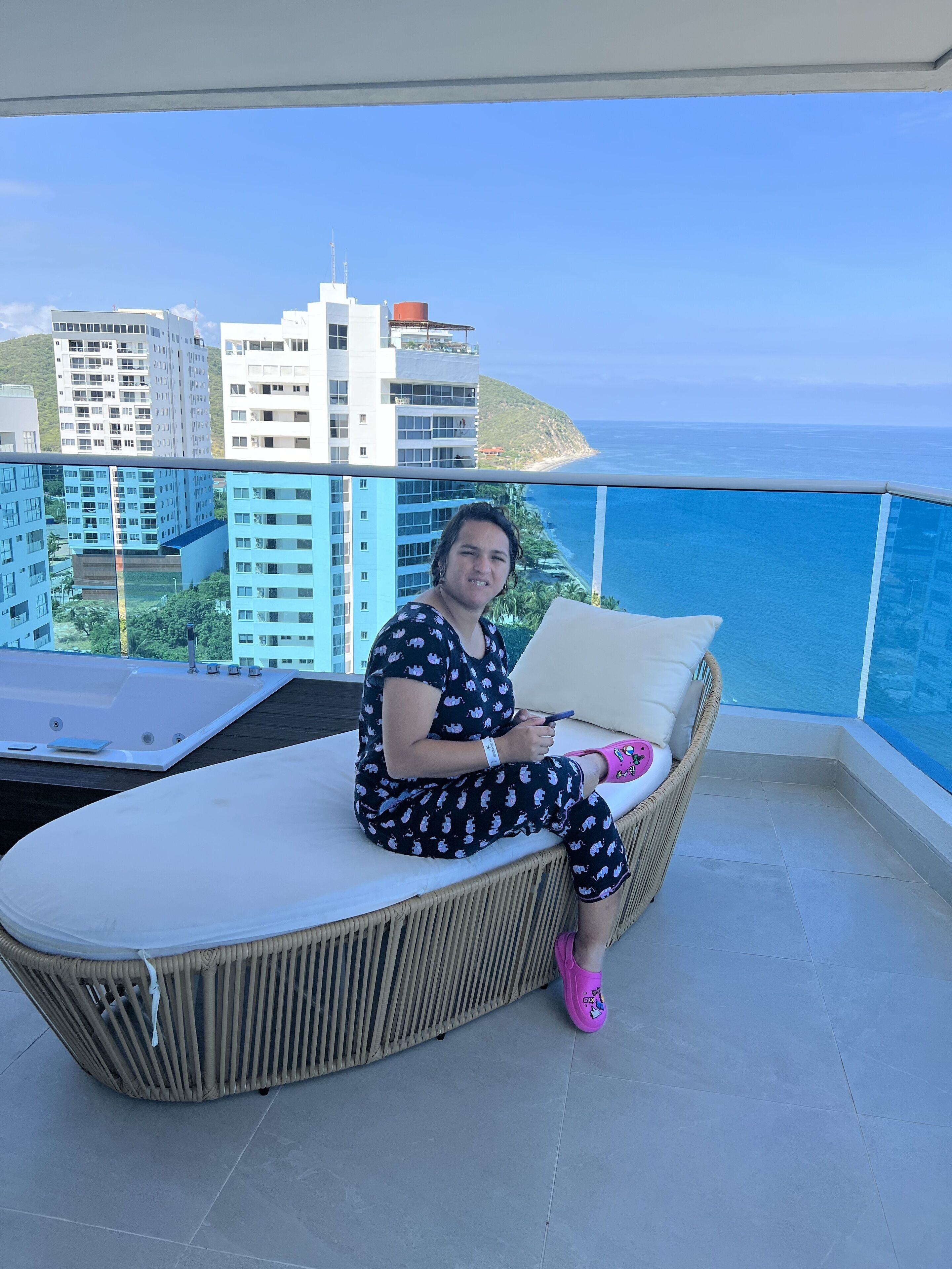Una persona seduta su una poltrona in vimini con un cuscino, che tiene in mano uno smartphone, su un balcone con vista sull'oceano e sugli edifici.
