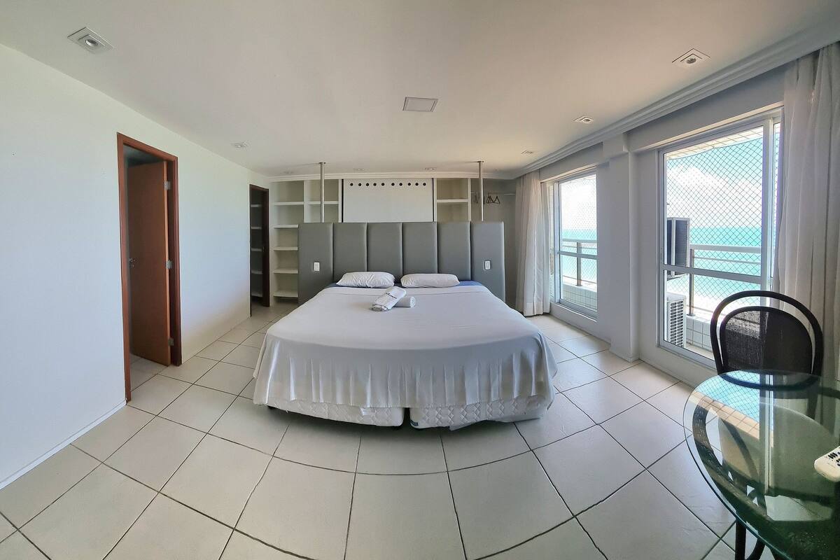 A modern bedroom with a large bed, built-in shelving, a glass table, and a balcony with a view.