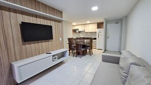 A modern living room with a flat-screen TV mounted on a wooden wall, a dining area with wooden chairs and a table, and a kitchen in the background. - Terraços do Atlântico By Solemar (Fortaleza)