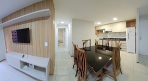 A modern living room with a flat-screen TV mounted on a wooden wall, a dining table with four chairs, and a kitchen area with white cabinets and appliances. - Terraços do Atlântico By Solemar (Fortaleza)