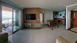 A spacious living area with a large TV mounted on a wooden panel wall, a dining table with chairs, and a balcony with a view of the sea. - Terraços do Atlântico By Solemar (Fortaleza)