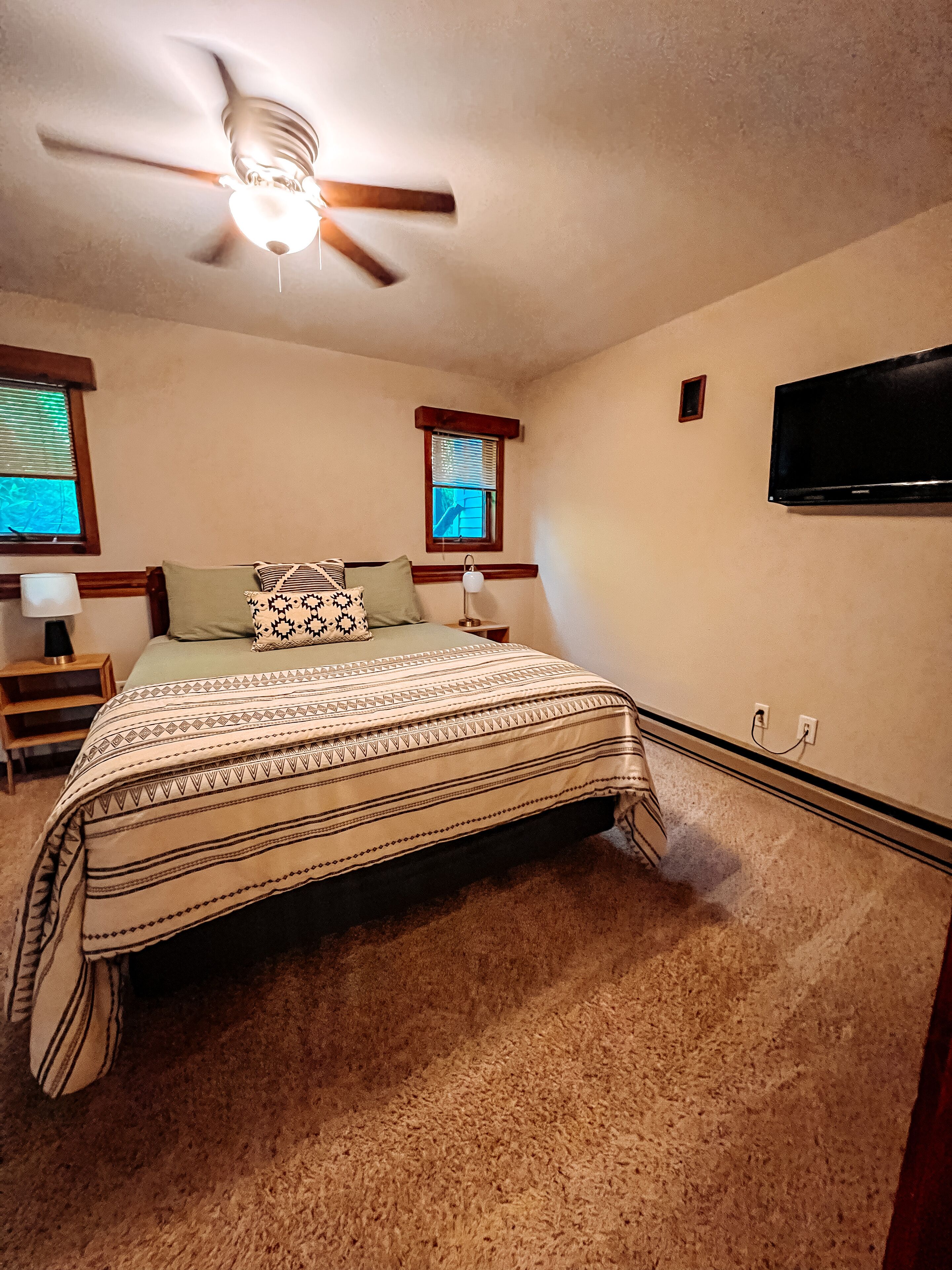 A bedroom with a bed, a ceiling fan, a TV mounted on the wall, and a window with blinds.
