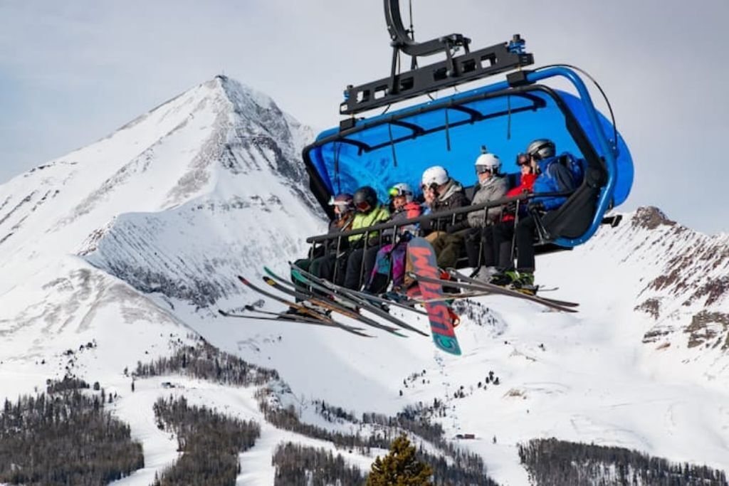 A ski lift carrying passengers with skis and snowboards, surrounded by snow-covered mountains.