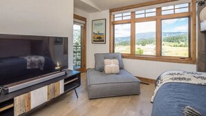 A bedroom with a bed, a TV, a grey chair, and a window with a mountain view. - Contemporary townhome in the heart of Town center (Big Sky)