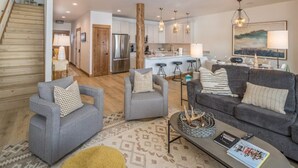 A spacious living area with a gray sofa set, a coffee table, and a patterned rug. A staircase leads to an upper level, and a kitchen with modern appliances is visible in the background. - Contemporary townhome in the heart of Town center (Big Sky)