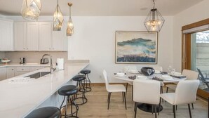 A modern kitchen with white cabinets, a large island with bar stools, pendant lighting, and a dining area with white chairs and a table set for four. - Contemporary townhome in the heart of Town center (Big Sky)
