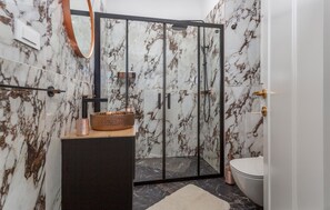 Moderni kylpyhuone, jossa on lasiseinällä varustettu suihku, marmoriseinät, puinen allaskaappi ja wc.