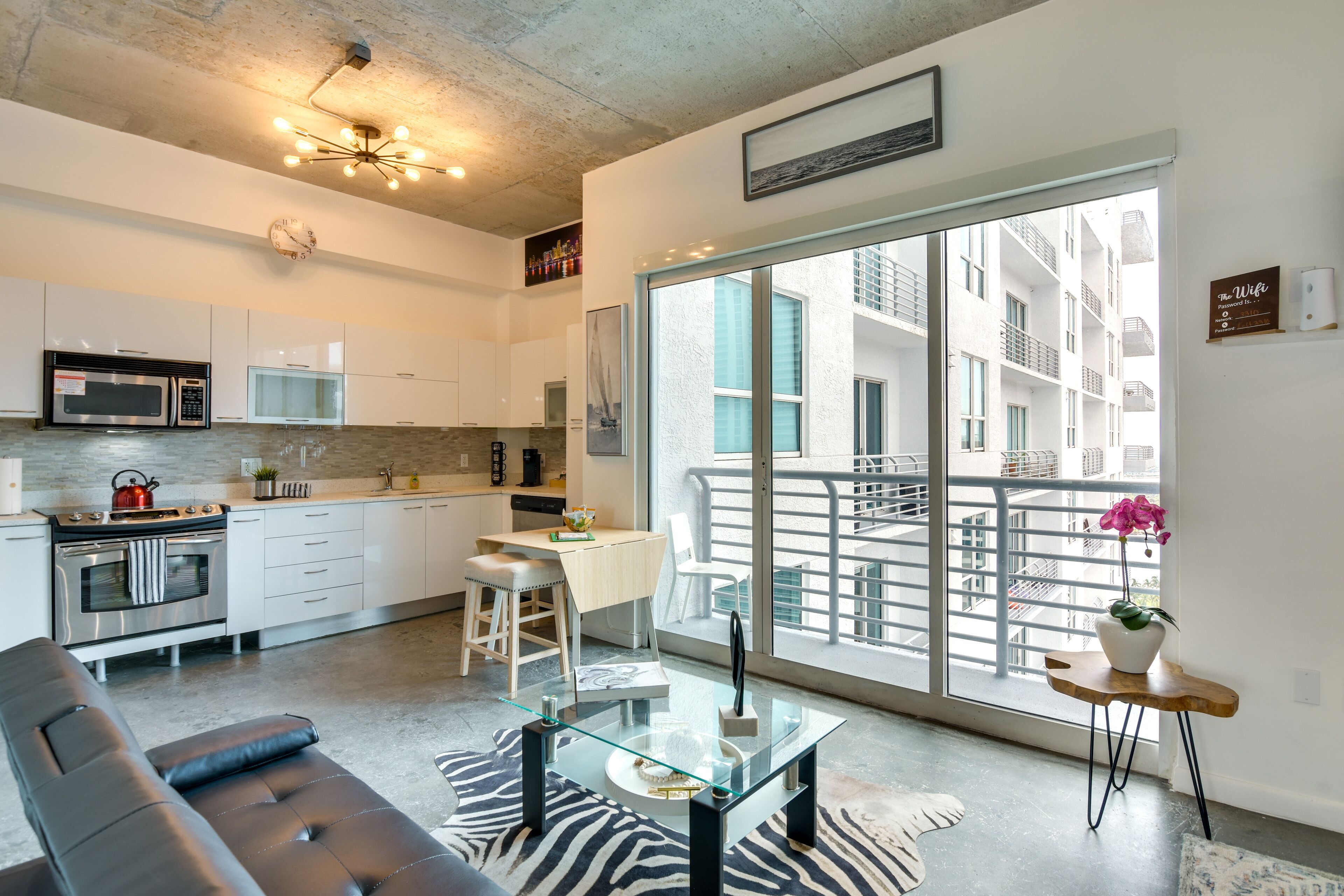 A modern apartment with a kitchen, dining area, and living room. It features a large sliding glass door to a balcony, a glass coffee table, and a black leather sofa.