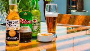 Una mesa con tres botellas de cerveza y un vaso, colocada al aire libre con cielo despejado y palmeras al fondo.