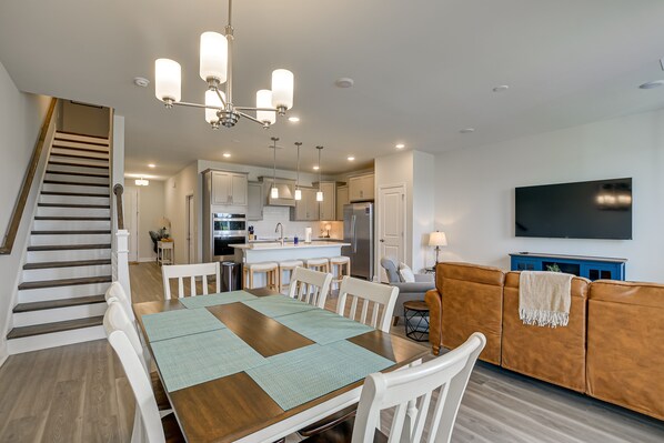 Una cocina moderna con una isla central, una zona de comedor con una mesa de madera y sillas blancas, y una sala de estar con un sofá marrón y un televisor montado en la pared.