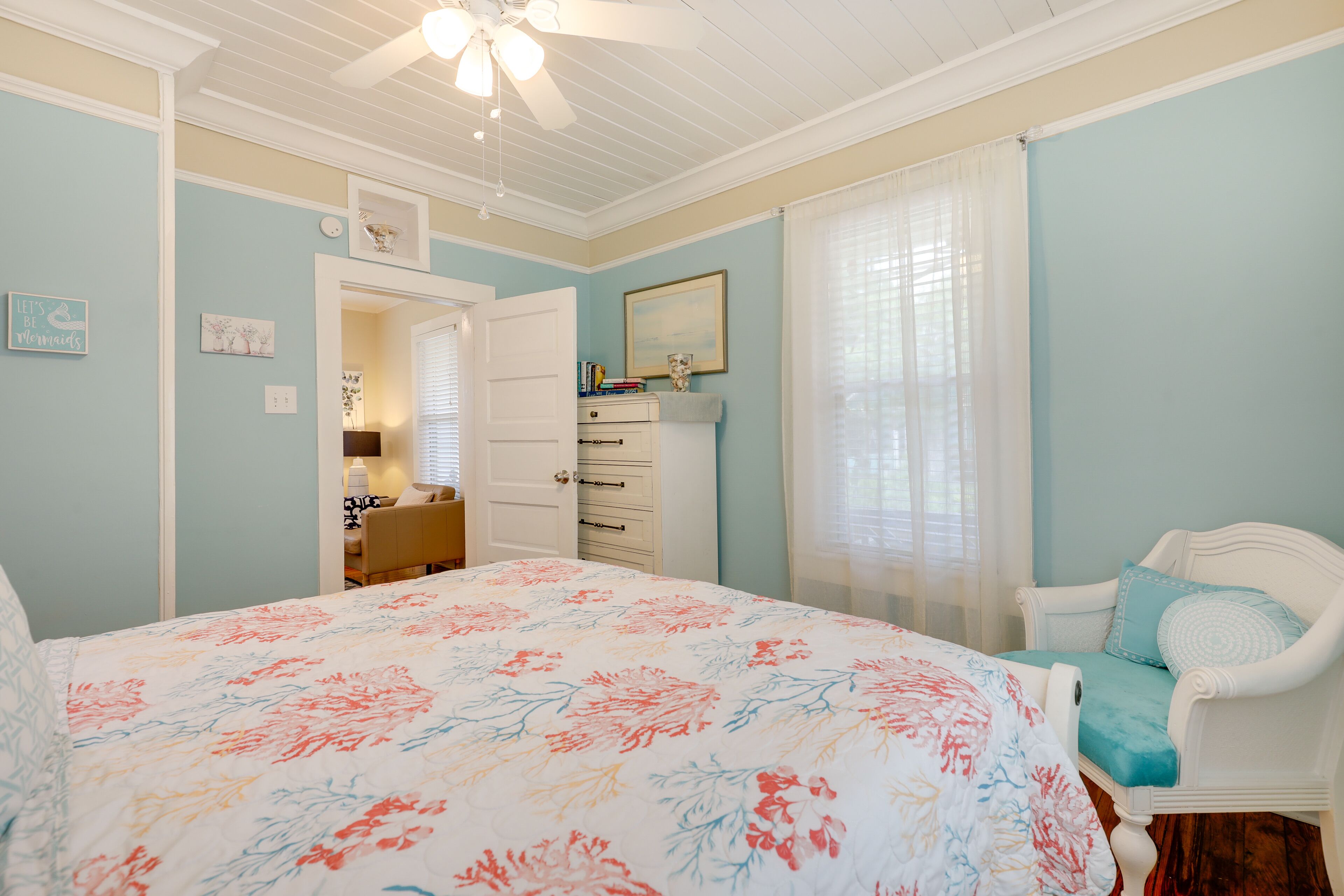 Ein Schlafzimmer mit geblümtem Bettüberwurf, einer weißen Kommode, einem Sessel mit blauer Sitzkissen und einem Deckenventilator.