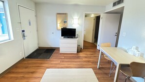 Une petite pièce bien éclairée, avec un plancher en bois, un bureau blanc avec une chaise, un téléviseur posé sur une commode et une porte donnant sur une autre pièce.