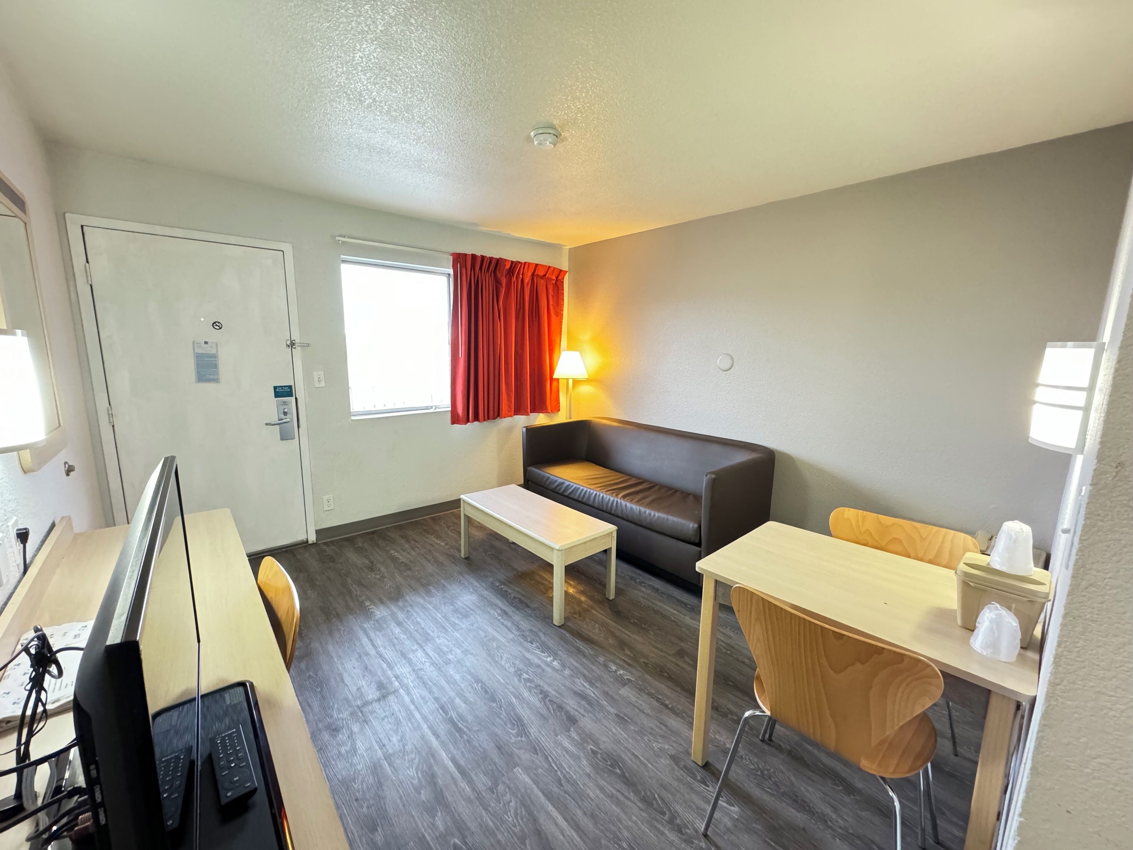 A compact hotel room with a single sofa, a small table, a desk with a computer, and a window with curtains.