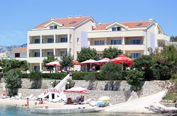 Ett hotell med flera balkonger, utomhusmöbler med parasoller och ett område vid stranden med solstolar.