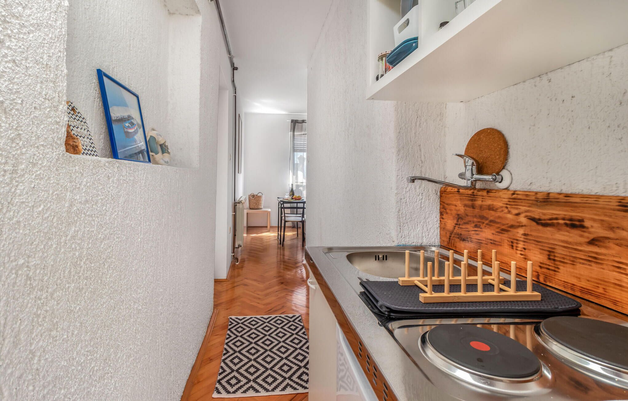 A narrow kitchen with a wooden countertop, a sink, and a small dining area in the background.
