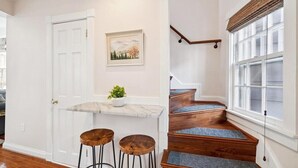 A staircase with wooden steps and blue carpet runners, a white door with a brass handle, and a small table with two stools and a potted plant. - Historic Home with Parking Steps From Capital & USNA (Annapolis)