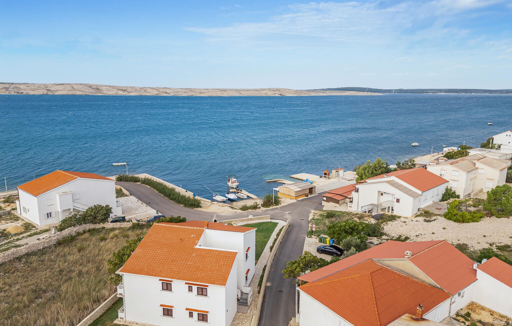 A coastal residential area with white buildings and red-tiled roofs, a marina with boats, and a clear view of the sea.