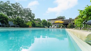 Ein Schwimmbecken mit Liegestühlen und einem gelben Sonnenschirm, umgeben von Grünflächen und einem Gebäude im Hintergrund.
