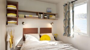Un dormitorio ordenado con cama, estantes con toallas dobladas, una mesita de noche con lámpara y una ventana con cortinas.