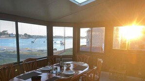 A dining area with a table set for four, large windows with blinds, and a view of the sea and boats.