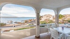 Un espacio al aire libre techado con vista al mar y barcos, una mesa con sillas, y un edificio al fondo.