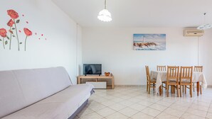 Una sala de estar con un sofá, un televisor sobre un mueble de madera, un cuadro de un faro y una mesa de comedor con sillas.