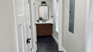 A hallway leading to a bathroom with a dark floor, white walls, and a wooden vanity with a mirror and sink.