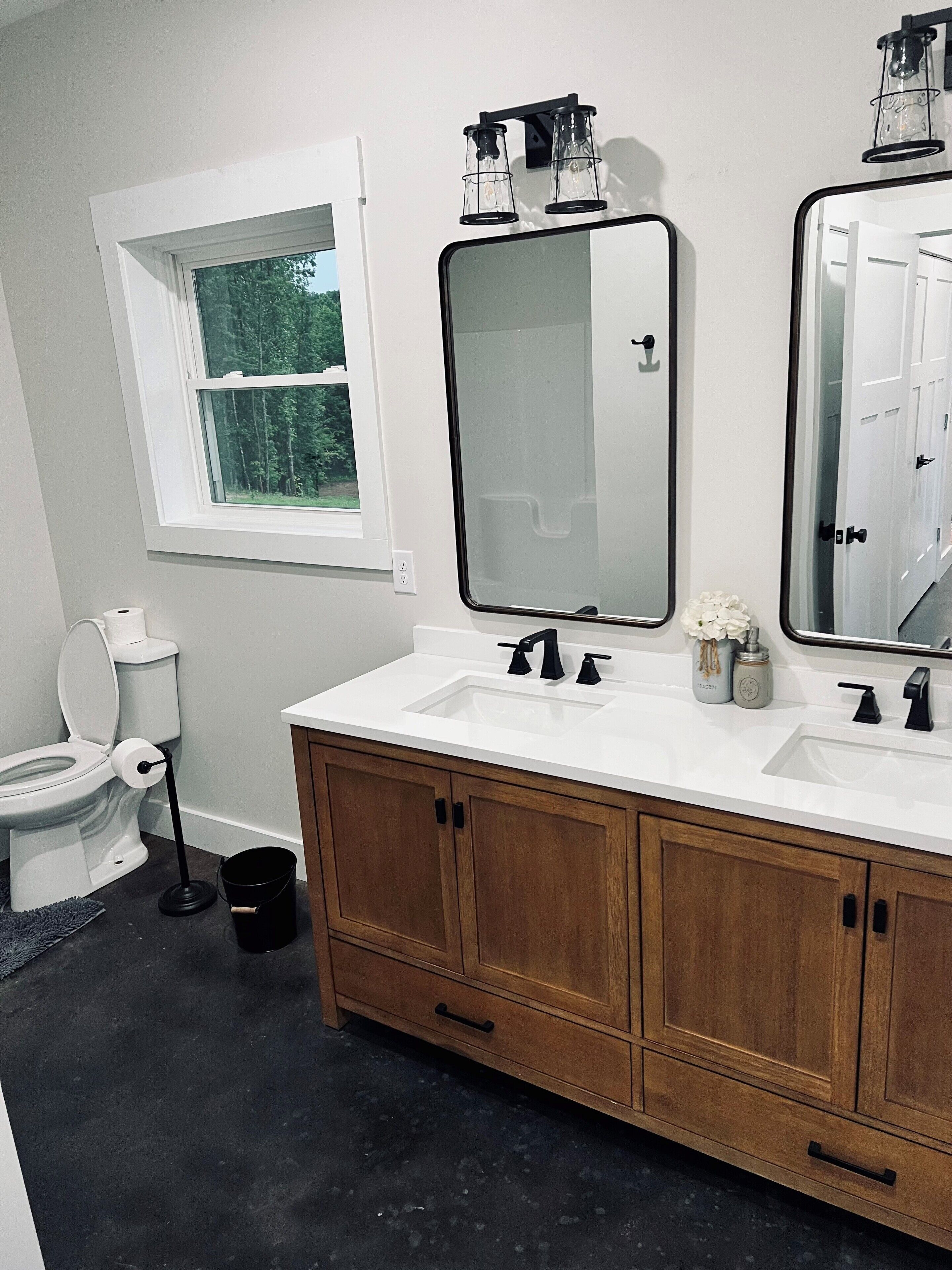 A bathroom with a white toilet, a sink with two faucets, a mirror, and a window with a view of trees.