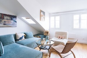 A modern living room with a blue sofa, a glass coffee table, and a wicker chair. There is a painting on the wall and a window with white frames. - Beautiful, Sun-kissed Studio—Faubourg Saint-Martin (Paris)