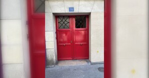 Una puerta roja con una placa de número, una pequeña ventana con diseño de cuadrícula y un muro de concreto.