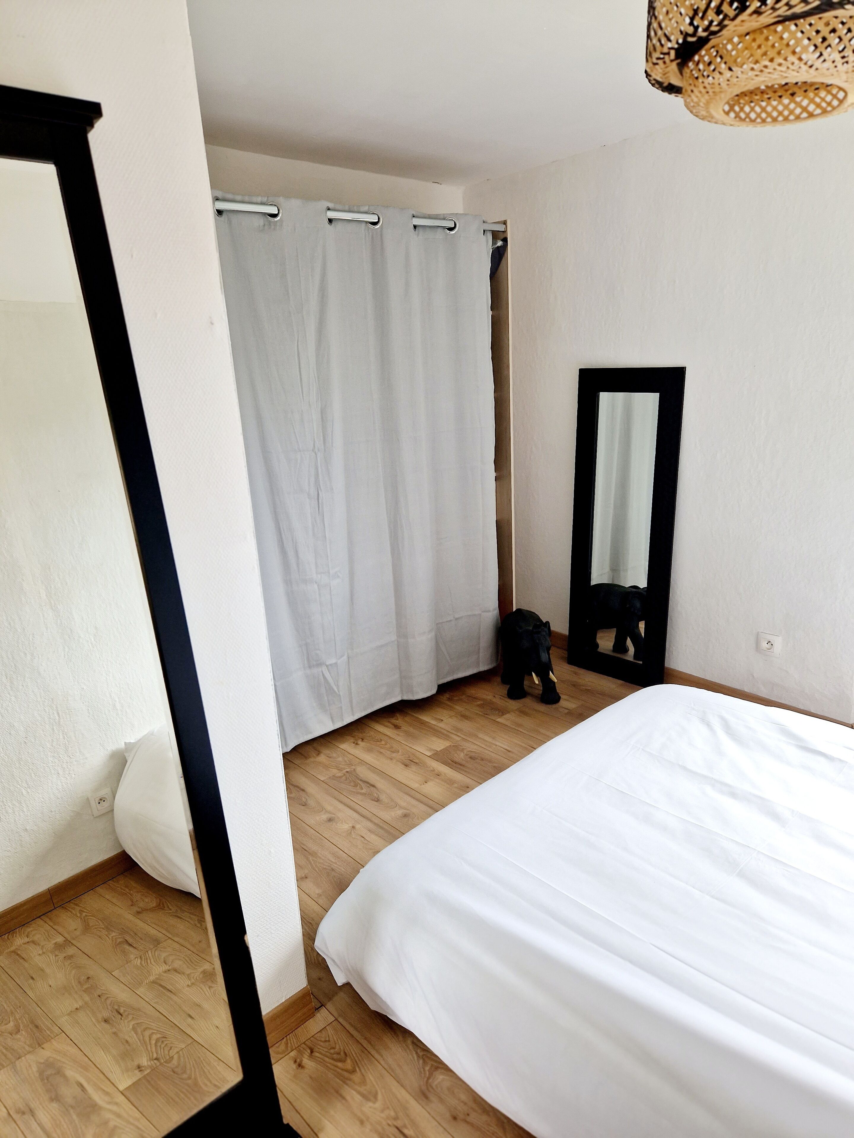 A bedroom with a bed, a wooden floor, a white curtain, a mirror, and a black cat.
