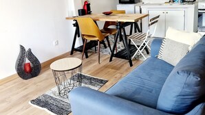 A modern living room with a blue sofa, wooden dining table, and yellow chairs. A kitchen with white cabinets and a microwave is visible in the background. - Maisonnette du Foirail, Proche Centre-ville Espalion. À 1 min des Berges du Lot! (Espalion)