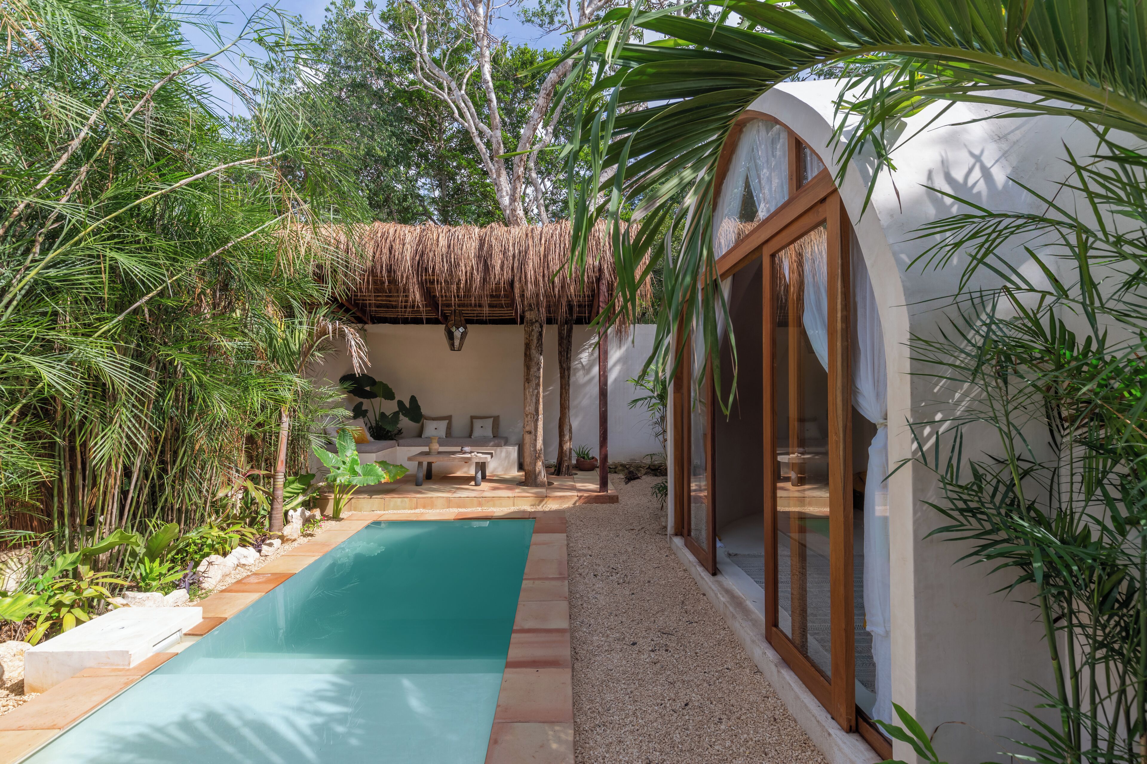 A pool area with a thatched roof structure, surrounded by tropical plants and a white building with large windows.