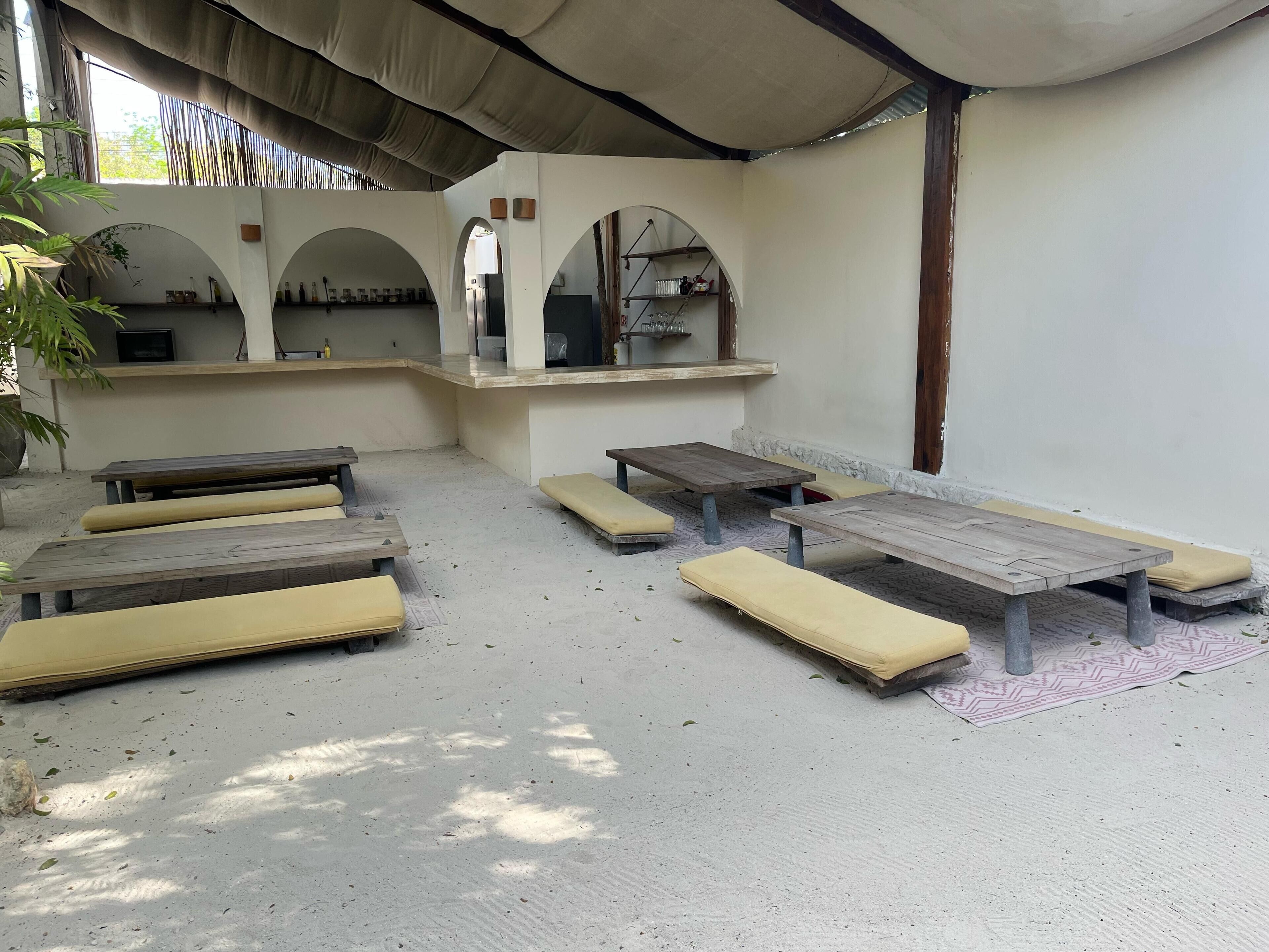 A covered outdoor seating area with benches and tables, a kitchen area with shelves, and a view of the outdoors.