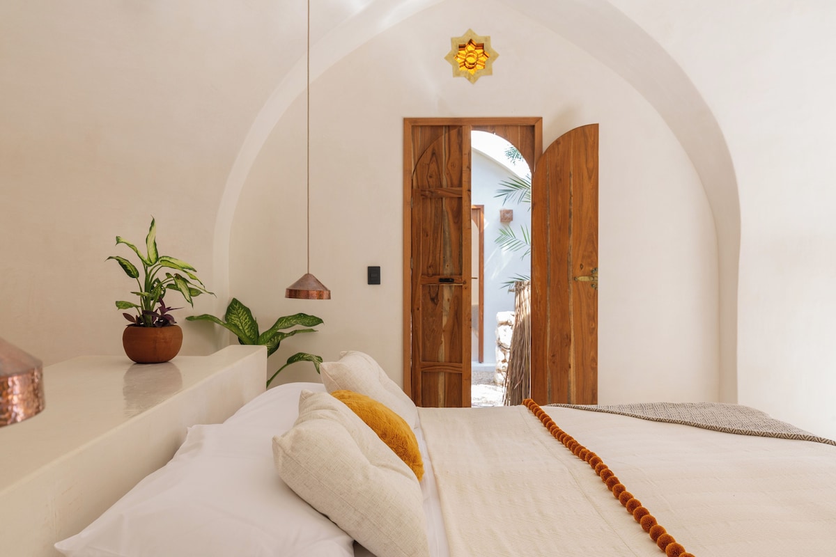 A bedroom with a bed, pillows, a hanging light, a wooden door, and a plant.