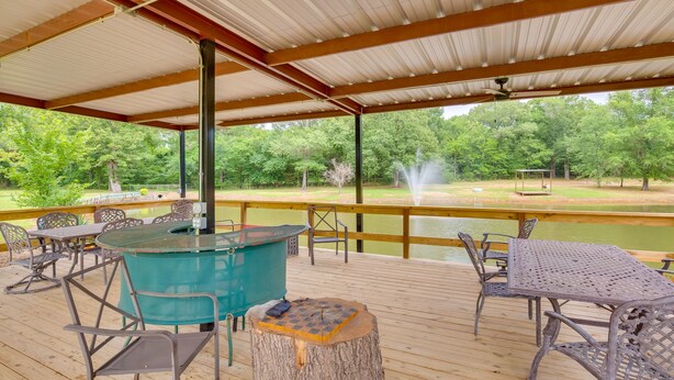 Une terrasse extérieure couverte, aménagée avec du mobilier en métal, un barbecue et offrant une vue sur un étang et des arbres.