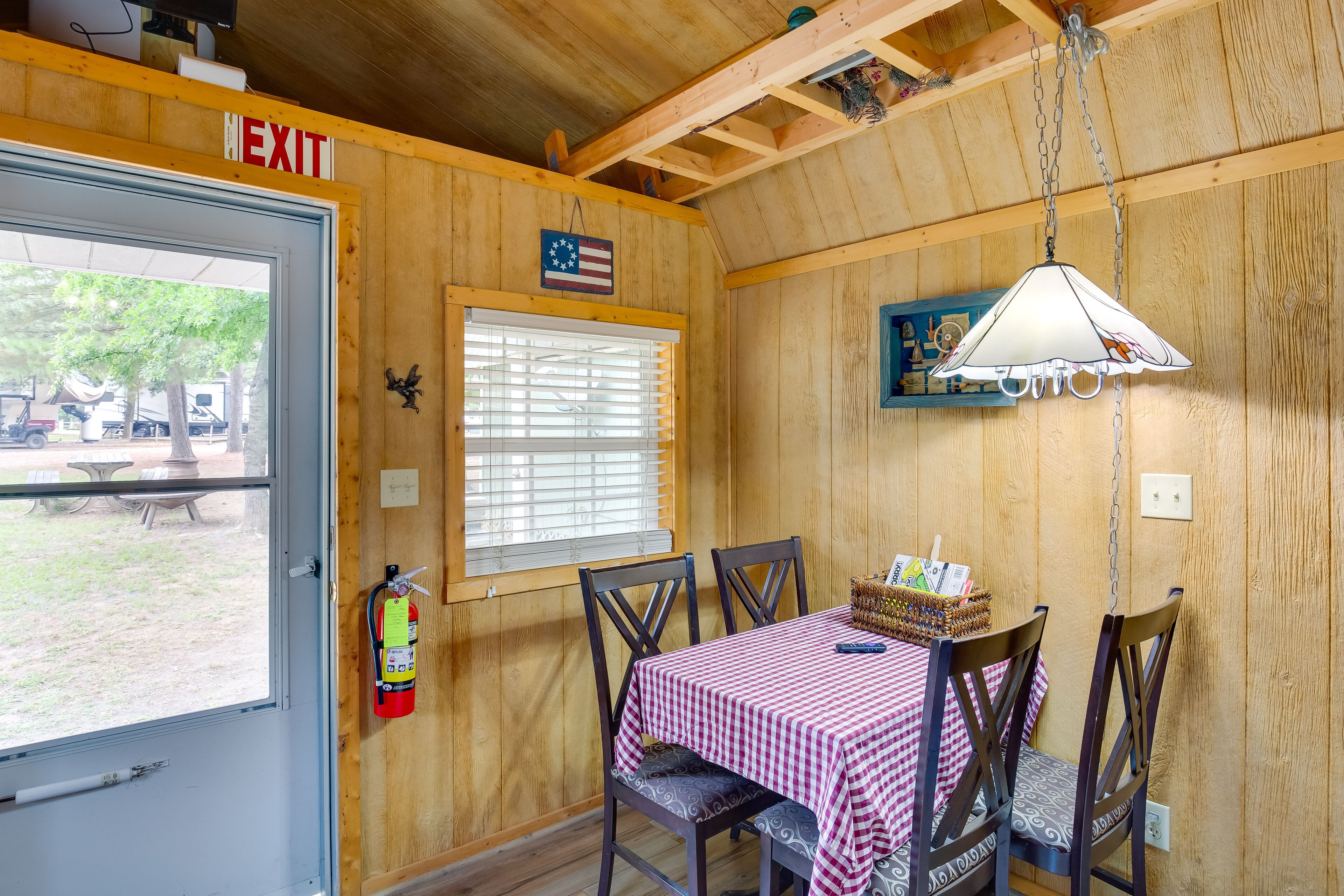 A wooden interior with a table covered in a checkered tablecloth, two chairs, a hanging lamp, a fire extinguisher, and an exit sign.