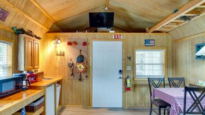 A compact kitchen with wooden cabinets, a microwave, a coffee maker, and a small refrigerator. There is a dining area with a table and chairs, and a television mounted on the ceiling. - Lake Fork Studio Cabin w/ Dock + Boat Ramp! (Emory)