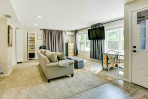 A living room with a sofa, ottoman, and a television. There is a window with curtains, a bookshelf, and a door. - Cozy Studio Stay in Liberty: Hike, Relax, Explore! (Liberty)